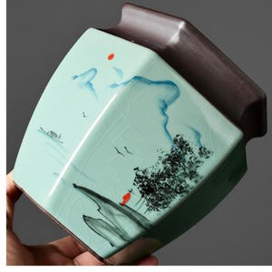 May include: A hexagonal ceramic tea canister featuring a light mint green crackle glaze finish. The exterior is decorated with a hand-painted landscape scene in black ink, depicting mountains, a tree, and a small figure in a red robe under a red sun.