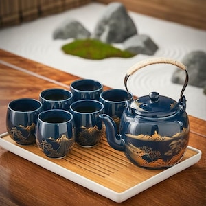 May include: A dark blue ceramic tea set comprising a teapot with a woven handle and five matching cups. Each piece features a gold-coloured landscape illustration of mountains and trees. The set is arranged on a rectangular bamboo tea tray.