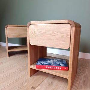 May include: Two light-colored wooden nightstands with rounded top edges, each featuring a single drawer and an open lower shelf. A book with a red spine and the white text KUKUCZKA is placed on the bottom shelf of the front nightstand.