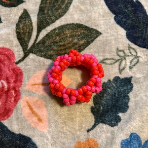 以下が含まれることがあります： 鮮やかなオレンジ色と明るいピンク色の小さなシードビーズで作られた円形のリング。ビーズは密度の高いテクスチャーのあるパターンで編み込まれており、繰り返される色の配列を持つ連続した丸みを帯びたバンドを形成しています。