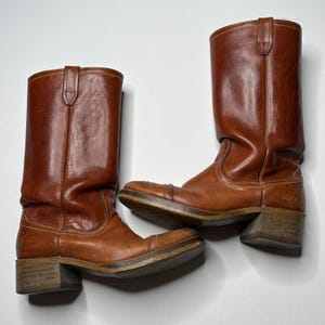 May include: A pair of mid-calf brown leather boots featuring a rounded toe, a stacked wooden heel, and visible stitching along the seams. The boots have a smooth finish with subtle natural variations in the leather texture and pull tabs at the top.