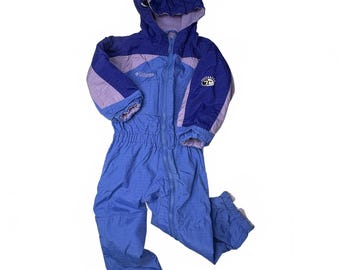 Puede incluir: Un traje de nieve de una pieza de la marca Columbia en tonos azul y lavanda. La prenda cuenta con una parte superior con capucha, una cremallera frontal completa y mangas largas con puños elásticos.