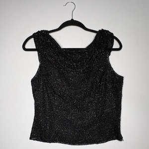 Op de afbeelding: Een mouwloze zwarte top met een volledig bewerkte textuur van kraaltjes en een gedrapeerde boothals. Het kledingstuk hangt aan een zwarte hanger en toont een getailleerd silhouet met ingewikkeld kralenwerk over het gehele vooroppervlak.