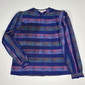 May include: A long-sleeved blouse featuring a horizontal plaid pattern in shades of royal blue, magenta, and teal. The garment has a round neckline, vertical pleats across the front bodice, and buttoned cuffs. A Michelle Stuart brand label is visible.