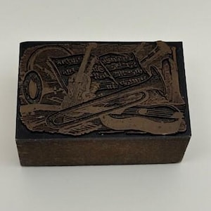 May include: A rectangular wooden printing block featuring a raised metal relief design of various musical instruments including a violin, a trombone, and a trumpet, alongside a sheet of musical notation, all set against a dark textured background.