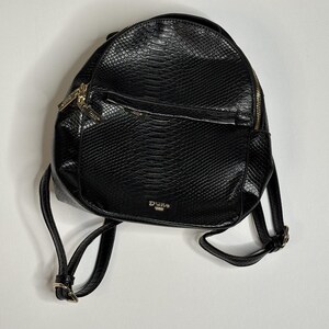 May include: A black backpack with a textured snakeskin pattern. The bag features gold-tone metal zips and hardware, including a front pocket zip pull and a small metallic logo plate at the bottom that reads Dune London.