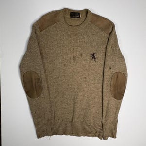May include: A tan crew neck sweater featuring brown suede shoulder patches and matching elbow patches. The knit garment displays a small dark brown embroidered lion logo on the chest and shows visible signs of wear, including small holes and fraying.