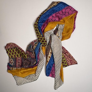 May include: A long scarf featuring a patchwork design of various fabric panels. The scarf displays a mix of patterns including black and white squares, floral motifs, geometric shapes, and solid sections in mustard yellow, bright blue, and pink.