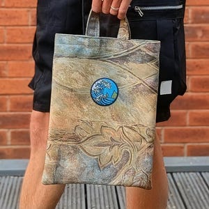 Könnte beinhalten: Eine rechteckige Stofftasche mit einer strukturierten, mehrfarbigen Oberfläche in erdigen Braun-, Beige- und Blautönen. Die Tasche zeigt ein geprägtes Blumenmuster und einen runden Aufnäher mit einer stilisierten blauen Meereswelle mit weißem Schaum.
