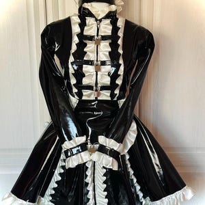 May include: A black and white glossy dress featuring long sleeves, a high collar, and a full skirt. The garment is adorned with white ruffles, three front metal padlocks on straps, and matching wrist cuffs with a padlock. A ruffled headpiece is included.