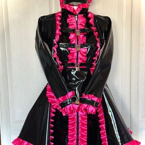 Könnte beinhalten: Ein langärmeliges Kleid in Schwarz und Pink mit hohem Kragen und passender Kopfbedeckung. Das Kleid hat glänzende schwarze Einsätze, leuchtend pinke Rüschen, einen zentralen Reißverschluss und schwarze Riemen mit silberfarbenen Vorhängeschlössern.