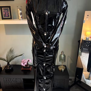 Könnte beinhalten: Ein langärmeliges, bodenlanges schwarzes Kleid aus einem glänzenden, lackartigen Material. Das Kleidungsstück verfügt über einen hohen Kragen und einen durchgehenden Frontreißverschluss, der mit mehreren kleinen silbernen Vorhängeschlössern gesichert ist.