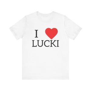 Könnte beinhalten: Ein weißes kurzärmeliges T-Shirt mit einem Grafikdruck auf der Brust. Das Design besteht aus dem schwarzen Buchstaben I, einem ausgefüllten roten Herzsymbol und dem Wort LUCKI in schwarzen Großbuchstaben direkt unter dem Herz.