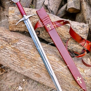 May include: A steel sword with a red leather-wrapped handle, a silver crossguard, and a pommel featuring a circular emblem. The blade is engraved with text. A matching red leather scabbard with gold cross accents and a leather strap is included.