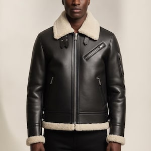 May include: A black leather jacket featuring a cream-colored shearling collar and cuffs. The jacket includes a central silver-tone zipper, two slanted zippered chest pockets, and two lower zippered pockets. Adjustable straps are at the collar.