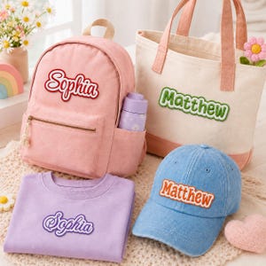 May include: A pink backpack with the name Sophia in red, a cream canvas tote bag with the name Matthew in green, a light blue denim baseball cap with the name Matthew in orange, and a lavender sweatshirt with the name Sophia in purple.