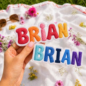 May include: Two embroidered fabric patches featuring the name BRIAN in bold, rounded, multi-colored block letters. The top patch has red, orange, pink, red, and yellow letters. The bottom patch has light blue, blue, dark blue, blue, and purple letters.
