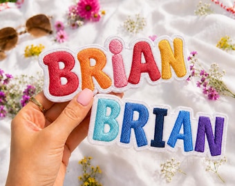 May include: Two embroidered fabric patches featuring the name BRIAN in bold, rounded, multi-colored block letters. The top patch has red, orange, pink, red, and yellow letters. The bottom patch has light blue, blue, dark blue, blue, and purple letters.