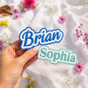 Puede incluir: Dos parches bordados con fondo blanco y bordes de colores. El parche superior muestra el nombre Brian en letras cursivas azules, mientras que el parche inferior muestra el nombre Sophia en letras cursivas de color turquesa con un borde a juego.