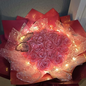 Könnte beinhalten: Ein Blumenstrauß aus hellrosa Satinband-Rosen, eingewickelt in dunkelrotes Papier und durchscheinendes, glitzerndes weißes Netzgewebe. Das Arrangement enthält ein Schmetterlingsornament auf der linken Seite und wird von kleinen LED-Lichtern beleuchtet.