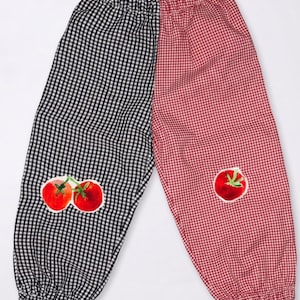 May include: Pants featuring a split design with one leg in black and white gingham and the other in red and white gingham. The black side has a pair of red tomato graphics, while the red side has a single red tomato graphic. The word batiko is printed.