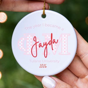 May include: A round white ceramic ornament featuring the text The year I became a, the name Jayda in red script, the Greek letters Phi Mu in a light pink checkered pattern, and the text Tulane University 2026 in red, suspended by a gold cord.