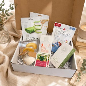 May include: A white cardboard gift box filled with various wellness items including a Saplaya aloe vera face mask, a pouch of Ginger Rescue lozenges, a bottle of Bio-Oil body lotion, a small handheld fan, Traditional Medicinals tea, and a glass jar.
