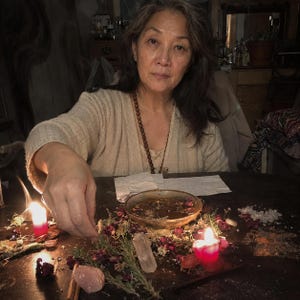 May include: A person wearing a beige cardigan and a brown beaded necklace sits at a dark table with a shallow bowl of liquid, scattered dried rose petals, rosemary sprigs, clear quartz crystals, a pink stone, and two lit red candles.