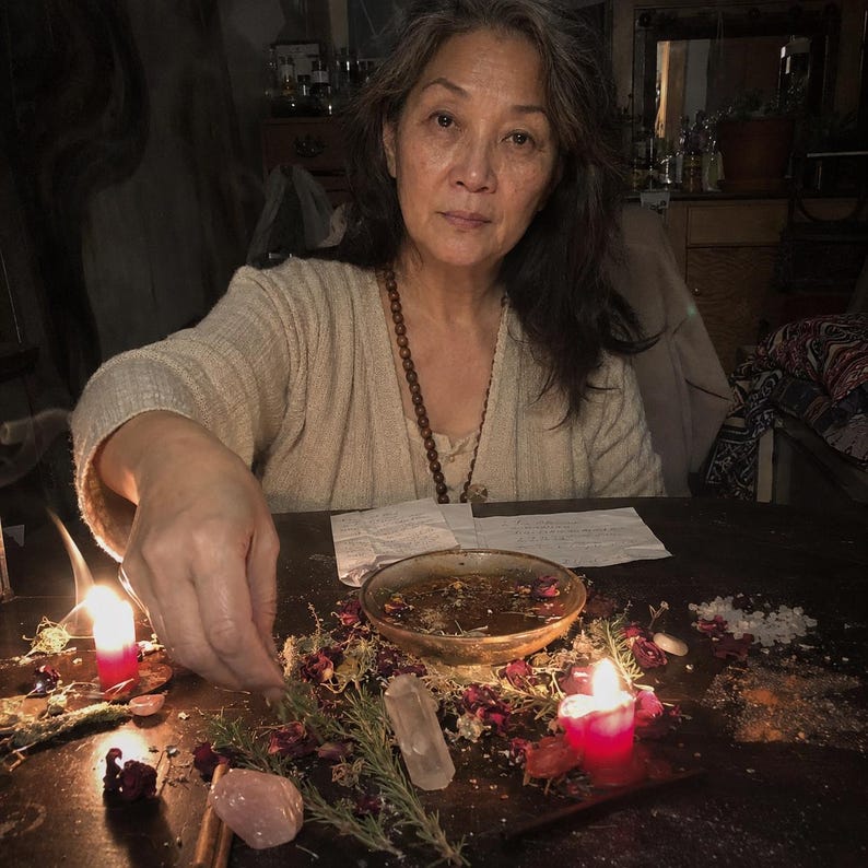 May include: A person wearing a beige cardigan and a brown beaded necklace sits at a dark table with a shallow bowl of liquid, scattered dried rose petals, rosemary sprigs, clear quartz crystals, a pink stone, and two lit red candles.