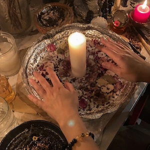 May include: A white pillar candle centered in a round aluminum foil tray filled with dried flower petals and bay leaves. The tray is surrounded by various items including a glass vase with purple flowers, a small Buddha statue, and a red candle.