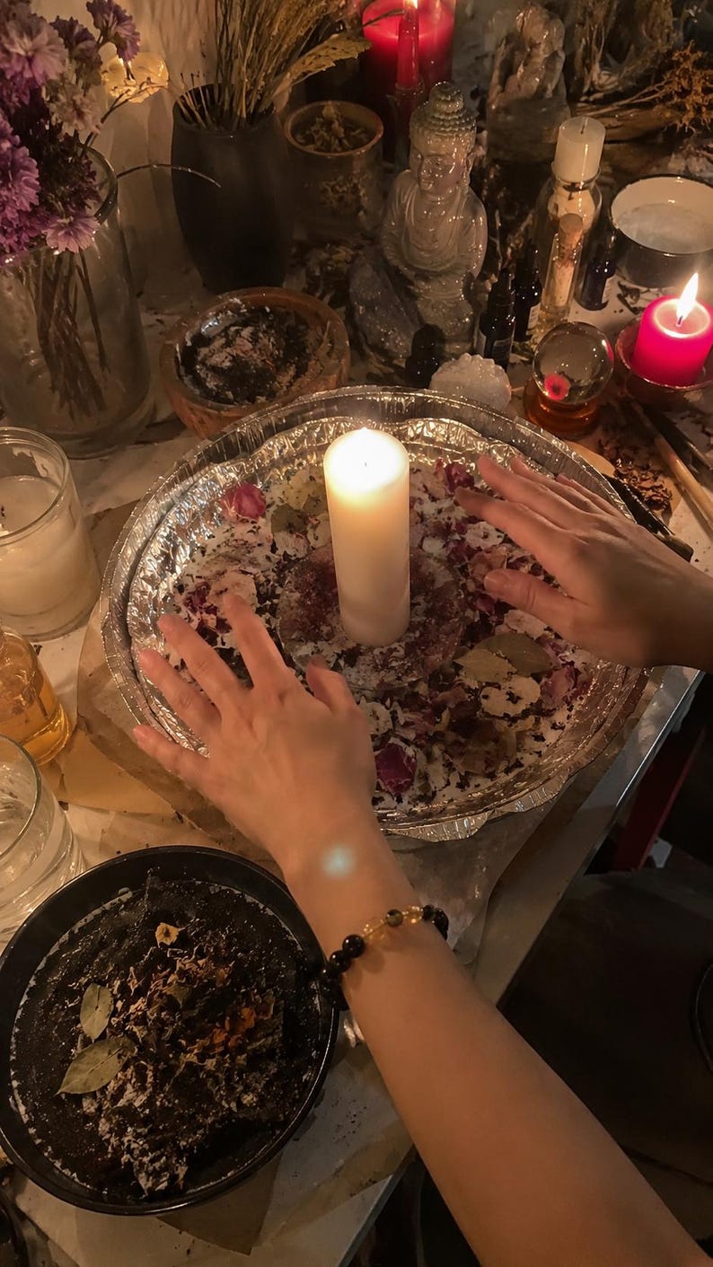 May include: A white pillar candle centered in a round aluminum foil tray filled with dried flower petals and bay leaves. The tray is surrounded by various items including a glass vase with purple flowers, a small Buddha statue, and a red candle.