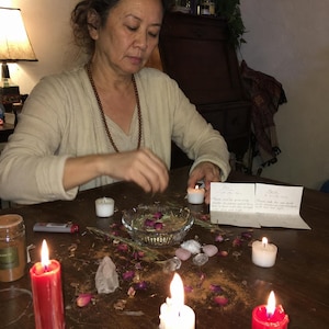 May include: A ritual setup featuring two red pillar candles and one white pillar candle, a glass bowl containing dried rosebuds and herbs, small clear and pink crystals, two small white tea light candles, and two handwritten note cards.