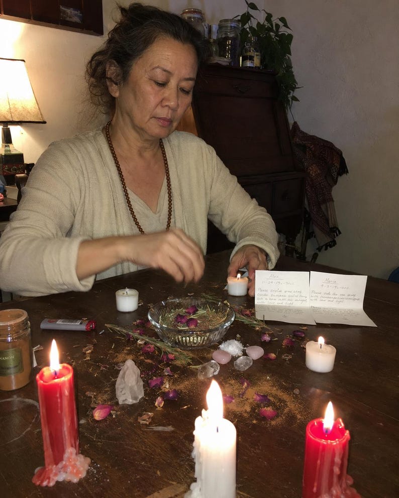 May include: A ritual setup featuring two red pillar candles and one white pillar candle, a glass bowl containing dried rosebuds and herbs, small clear and pink crystals, two small white tea light candles, and two handwritten note cards.