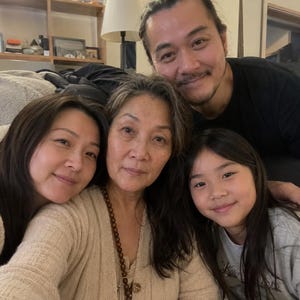 May include: A group of four people posing together for a photo. The group includes a man with dark hair, an older woman with dark hair wearing a beige cardigan and wooden bead necklace, and two younger individuals with long dark hair.