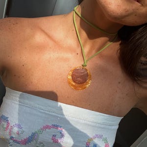 Puede incluir: Un collar con un cordón verde claro y un colgante circular. El colgante consta de un disco más grande de color ámbar translúcido con una superficie texturizada, superpuesto con una pieza circular más pequeña de color marrón rojizo oscuro.