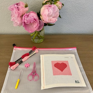 May include: A needlepoint kit featuring a square canvas with a stitched red heart on a pink background and the letters RS in the corner. Includes a clear zippered pouch, two skeins of red and pink embroidery floss, pink scissors, and a yellow needle tool.