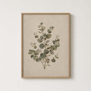 May include: A framed wall art print featuring a botanical illustration of a eucalyptus branch with muted green, rounded leaves. The artwork is set against a textured, light beige background and enclosed in a simple, light-toned wooden frame.