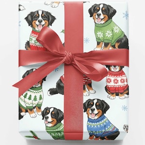 May include: A rectangular gift box wrapped in white paper featuring a repeating pattern of Bernese Mountain Dogs wearing colorful holiday sweaters, tied with a wide red satin ribbon and a bow centered on the front of the package.