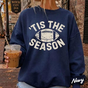 Puede incluir: Una sudadera azul marino de cuello redondo con un estampado gráfico blanco desgastado de un balón de fútbol americano centrado entre el texto 'TIS THE SEASON. El diseño está impreso con una tipografía de estilo vintage en el pecho.