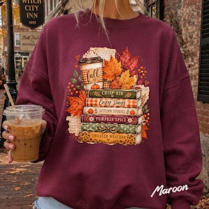 May include: A maroon crewneck sweatshirt featuring a graphic print of a coffee cup labeled fall, surrounded by autumn leaves and a stack of five books titled Crisp Air, Cozy Days, Autumn Stories, Pumpkin Spice, and Sweater Weather.