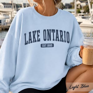 May include: A light blue crewneck sweatshirt featuring the dark blue text LAKE ONTARIO in a distressed font, with the text EST 1660 enclosed in a rectangular box underneath. The garment has long sleeves and a ribbed collar, cuffs, and hem.