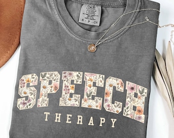 May include: A charcoal gray short-sleeve t-shirt with a crew neckline. The word SPEECH is printed in large block letters filled with a floral pattern of daisies and wildflowers. The word THERAPY is printed below in smaller, plain beige letters.