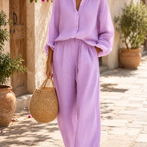 May include: A lavender long-sleeved button-down shirt with a collar and matching wide-leg trousers. The outfit is paired with a round, woven straw handbag and gold-toned hoop earrings, creating a coordinated set in a light purple hue.