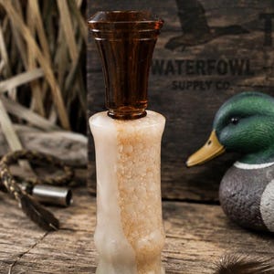 May include: A duck call featuring a translucent dark brown acrylic barrel and a textured, cream-coloured resin insert with a bubbly, organic surface pattern. The call has a cylindrical body that tapers slightly towards the centre and a flared top.