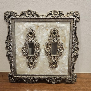 May include: A double light switch plate cover featuring an ornate, antiqued gold-tone metal frame with floral detailing. The center panel is made of a cream-colored, iridescent mother-of-pearl material, and two matching metal switch plates are mounted on top.