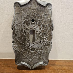 May include: A rectangular light switch wall plate with a decorative, scalloped edge. The surface features a dark grey and silver crackled, stone-like texture with a metallic finish. It includes two screw holes and a central rectangular toggle cutout.
