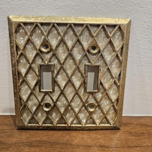 May include: A double light switch wall plate featuring a gold-toned metal frame with a diamond lattice pattern overlaying a textured, pearlescent white background. The rectangular plate includes two vertical rectangular cutouts for switches.