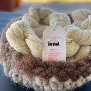 May include: A close-up of a skein of natural white alpaca yarn, with a brown and white yarn skein in the background. A white tag with pink lettering reads "Gulf Breeze Alpaca & Sheep Ranch" and includes the address "200 Jakeview Drive, Lake City, TX 78366".