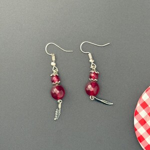 Pode incluir: Um par de brincos pendentes com ganchos prateados, cada um adornado com uma pequena conta vermelha facetada, uma conta vermelha facetada maior e um pingente de pena de metal prateado na base, conectados por calotas decorativas prateadas.