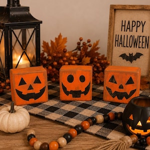 May include: Three orange wooden blocks with black painted jack-o'-lantern faces, a black metal lantern, a small white pumpkin, a black pumpkin-shaped candle holder, a wooden bead garland with a tassel, and a sign that reads HAPPY HALLOWEEN.